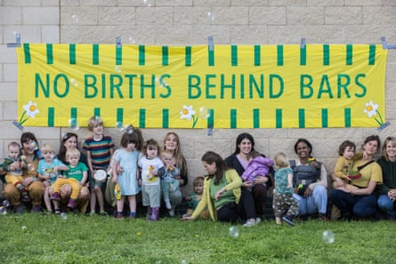 A group of women with small children crouch on the grass in front of a banner on a wall reading ‘No births behind bars’