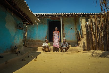 Indian woman with cowry shell necklaces and bracelets poses with an old man and old woman who are squatting in the yard of a single-storey home with tin roof