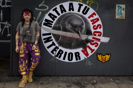 A young woman with streaks of red in her hair and tattoos leans against a wall with a picture of a crow with a knife in its beak