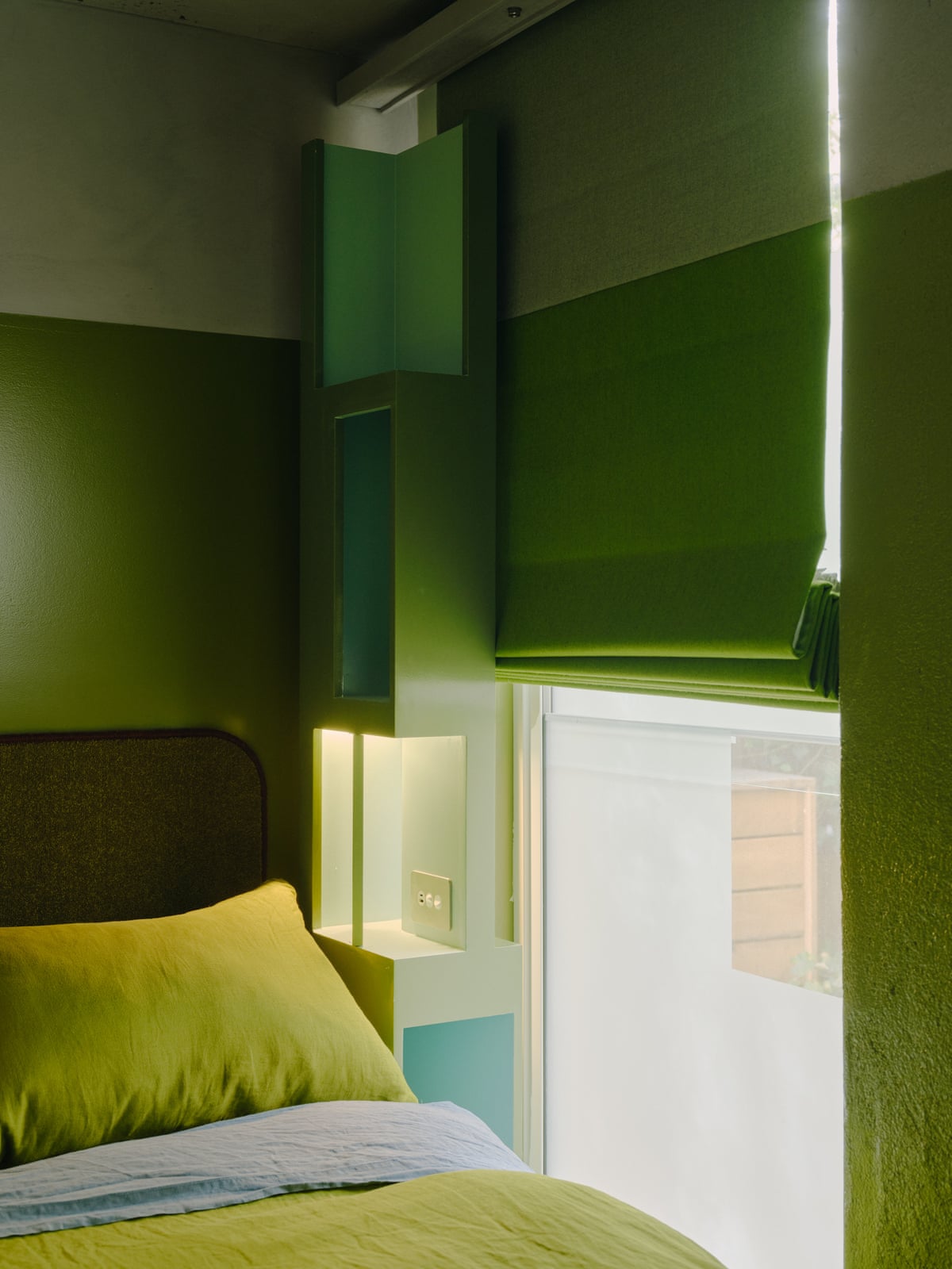 A green roman blind hangs beside an illuminated shelving unit above a bed