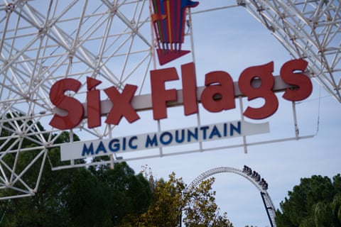 Biển hiệu lối vào lớn của Six Flags Magic Mountain uốn cong trên những tán cây, phía dưới là vòng xoay của tàu lượn siêu tốc.