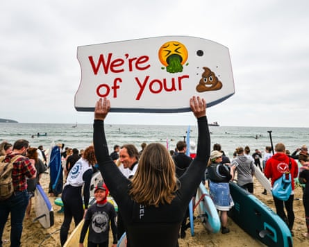 A group of surfers on a beach, one of them hold up a board reading ‘we’re sick of your poo’ illustrated by emojis