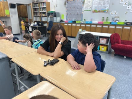 An adult and a child sit at low desks next to each other in a classroom looking at each other, both with an arm on the desk and left hand on the face.
