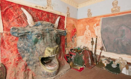 A room with a large minotaur head sculpted on to a red wall with the fireplace as the mouth. Busts of ancient figures are painted around the top of the walls, with ancient figures also painted on an orange wall. What appear to be Roman costumes lie on the floor