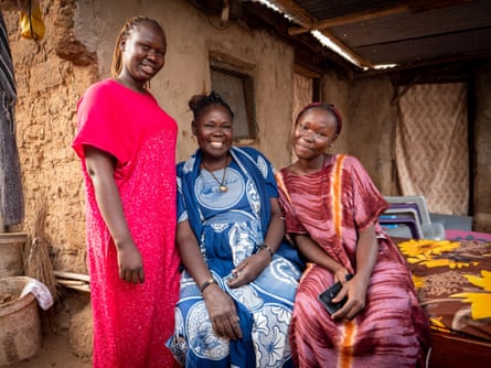 A mother sits between her two daughters outside a mud-walled house