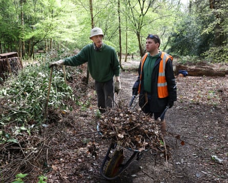 Two people move green material in a wood; one is wearing a high vis and pushing a wheelbarrow