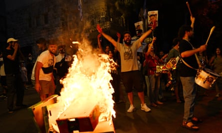  a group of young men are seen behind a fire in the street, some waving their arms and shouting; one man is banging a drum