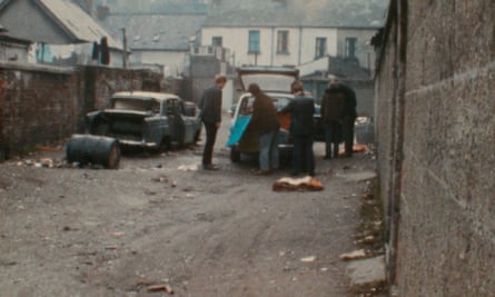 Two people lean into a car boot, with two others standing by, down an alley with worn-looking houses at the end