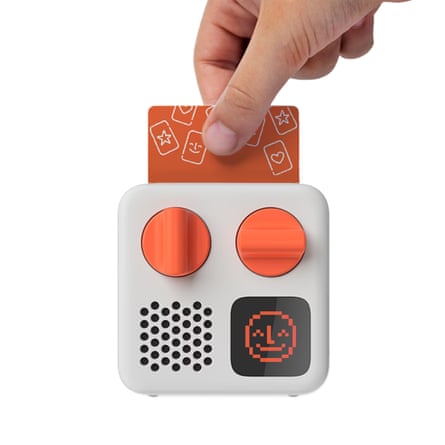 A hand inserts an orange card into the top of a Yoto player with dials and a smiling pixel face