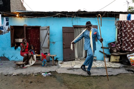 Photograph of a very dapper man dancing outside a blue house while a woman watches on