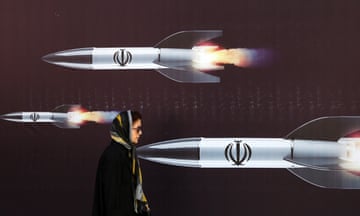 A woman walks past a poster of three missile-type weapons firing from right to left