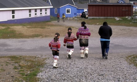 An Inuit mother and girls wearing the traditional costume embroidered with colored pearls walk on a dirt path away from the camera toward a blue building in Qeqertarsuaq, Greenland
