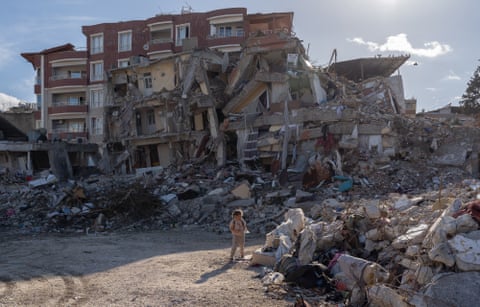 A very young girl stands on a patch of waste ground in front of a largely destroyed apartment block