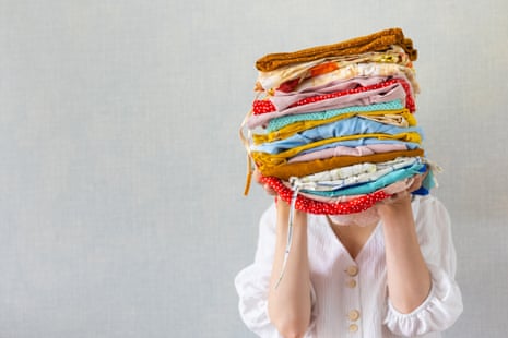 A woman wearing a white blouse holds a pile of folded, colourful clothes in her hands, covering her face.