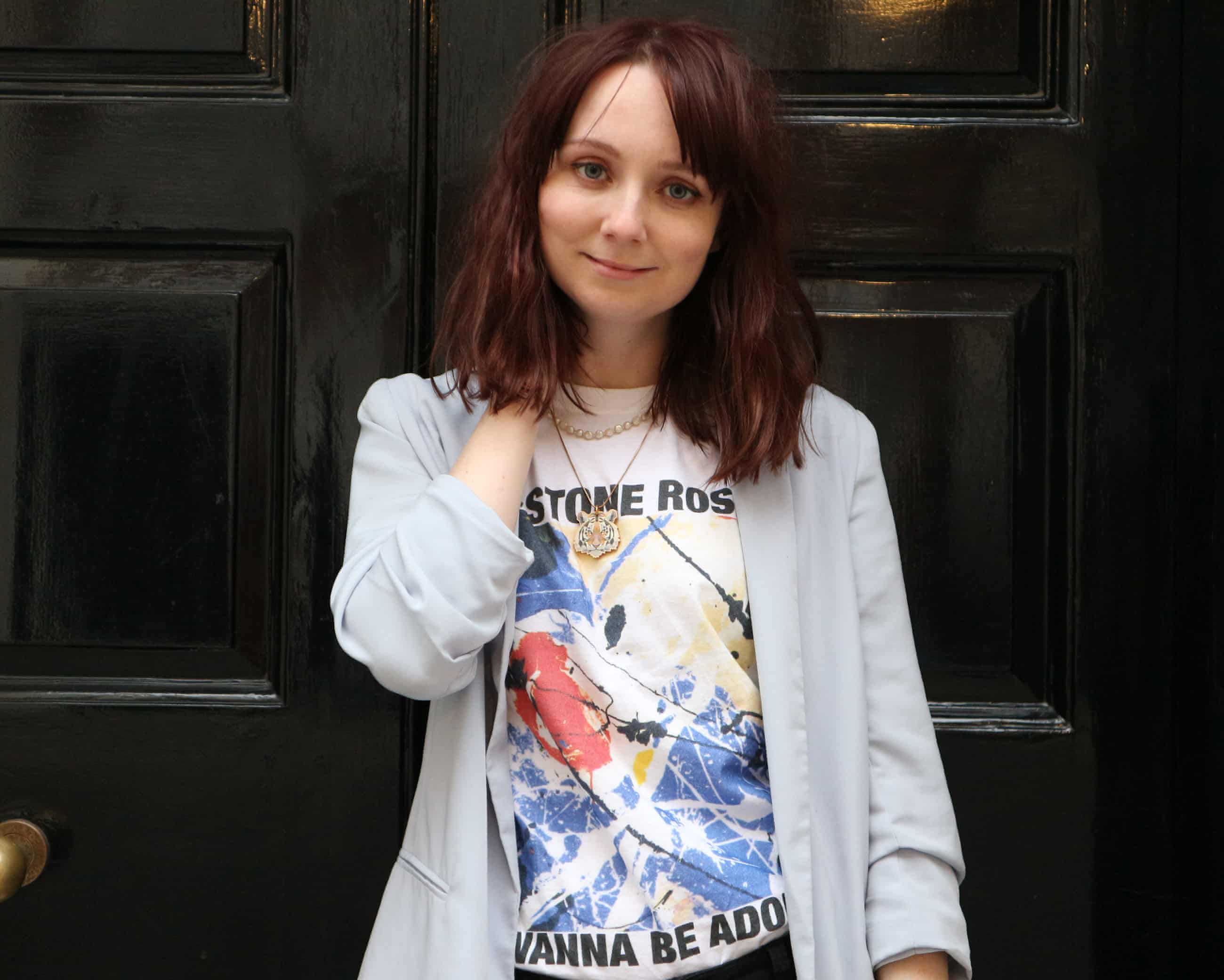 close up of Claire Eastham wearing white jacket and white Stone Roses T-shirt with shoulder length brown hair in front of a black front door