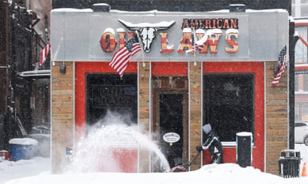 A crest of snow blows from a white skid in front of a red, gray and brick building with a longhorn skull logo.
