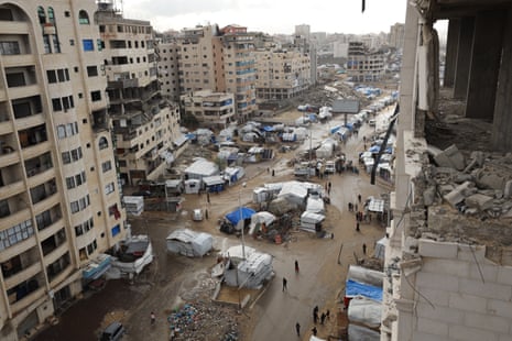 A view up a wide road flanked by bombed out apartment buildings, around the base of which are tarpaulin shelters, which also cover teh road's central reservation.
