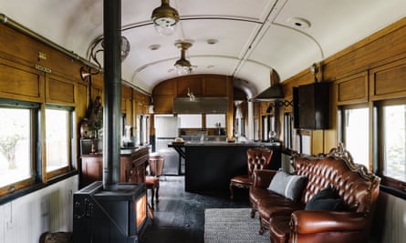 The interior of the train carriage with a studded leather sofa and wood-burning stove in the foreground, a stainless steel kitchen at the far end and wood panelling throughout