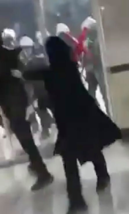 Blurred video screengrab shows a woman in black headscarf and robe who appears to be turning to run as what look like riot police in helmets line up at glass doors behind her.