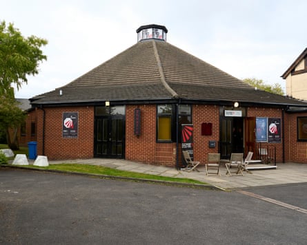 A single-storey brick building with a steep roof and skylight in the centre. It has placards advertising the plays and wooden chairs placed by the door.