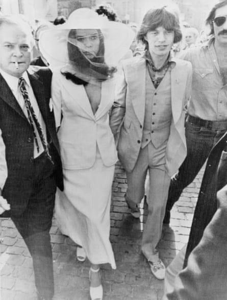 A woman in a skirt suit and a veiled wide-brimmed hat walking alongside a man in a suit as they are surrounded by other men.