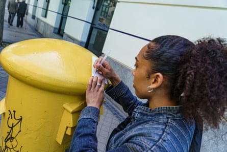 A woman leans on a yellow letterbox as she writes on a small piece of paper.