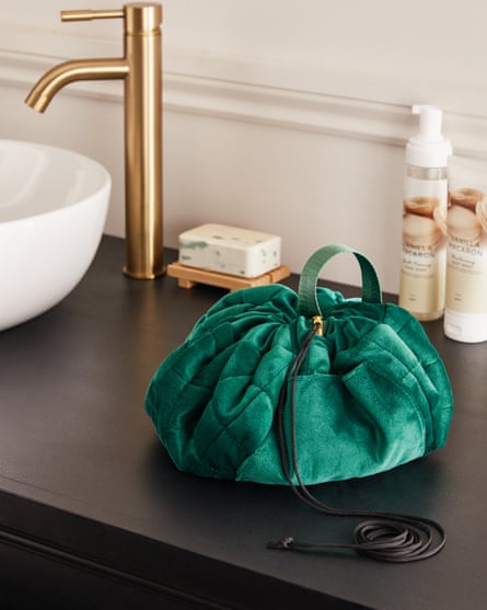 A green velvet drawstring makeup bag on a dark wood bathroom sink counter next to a white sink with a gold tap, which also features a bar of soap and two tubes of skincare