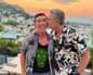 A man kisses his partner on the cheek on an Athens rooftop terrace at sunset, with white buildings and hills behind