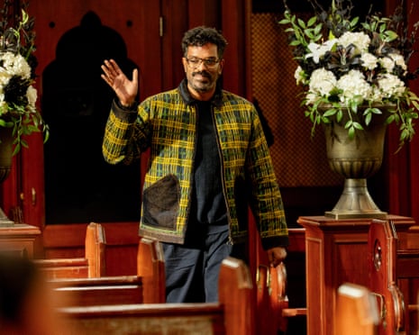 Romesh Ranganathan in a plaid jacket waves while walking through wooden church pews