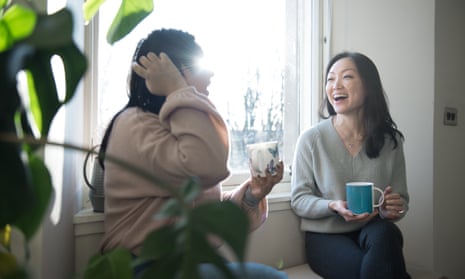 Two women chat and laugh while having a cup of tea sitting next to a window.