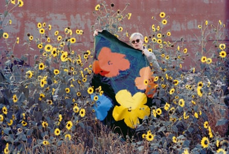 Andy Warhol in a field of flowers holding one of his flower paintings, against a wall with peeling paint