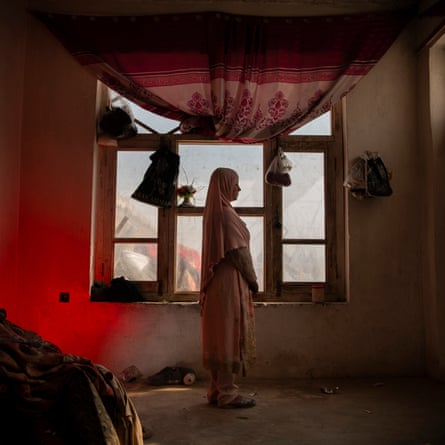 A woman wearing a headscarf stands in a room next to the window