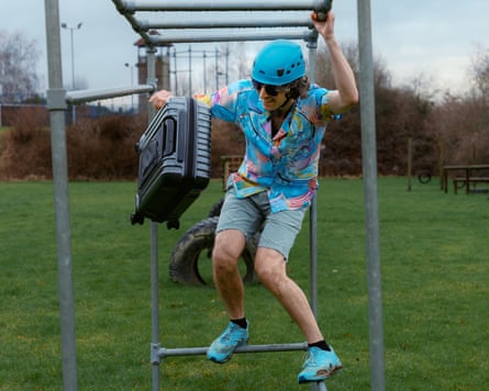 The writer Pete Wise is working his way through a climbing frame with a suitcase in tow! He wears a bright coloured shirt and has knee-length shorts.
