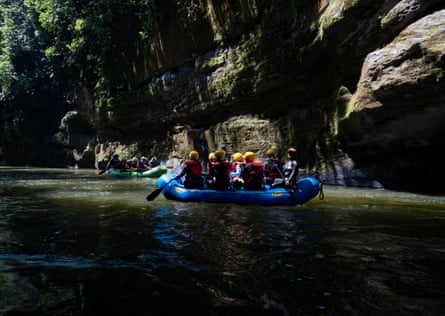 A few bright blue river rafts full of people wearing life vests and holding oars, on a green river alongside a rocky cliff the expands high beyond the frame.