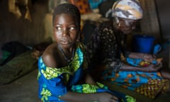 An apprehensive-looking girl and an older woman in traditional dress sit in a hut