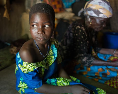 An apprehensive-looking girl and an older woman in traditional dress sit in a hut