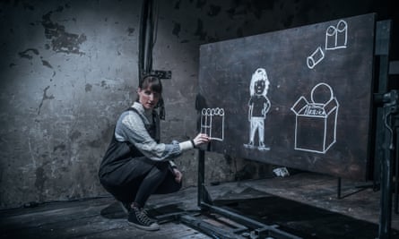 a woman crouches near a blackboard in a dark, forbidding room, in a scene from the show Somewhere Else by Slovenia’s Ljubljana Puppet theatre.