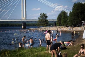 People stand, sit and bathe at a beach on the river’s edge, with a road bridge in the background