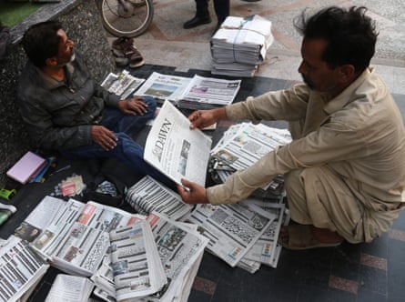 Man crouched reading a front page amid a pile of newspapers and vendor seated