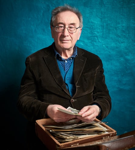 George Szirtes, in a shirt, cardigan, glasses and jacket, holding the typewriter case and photographs he carried when fleeing Hungary