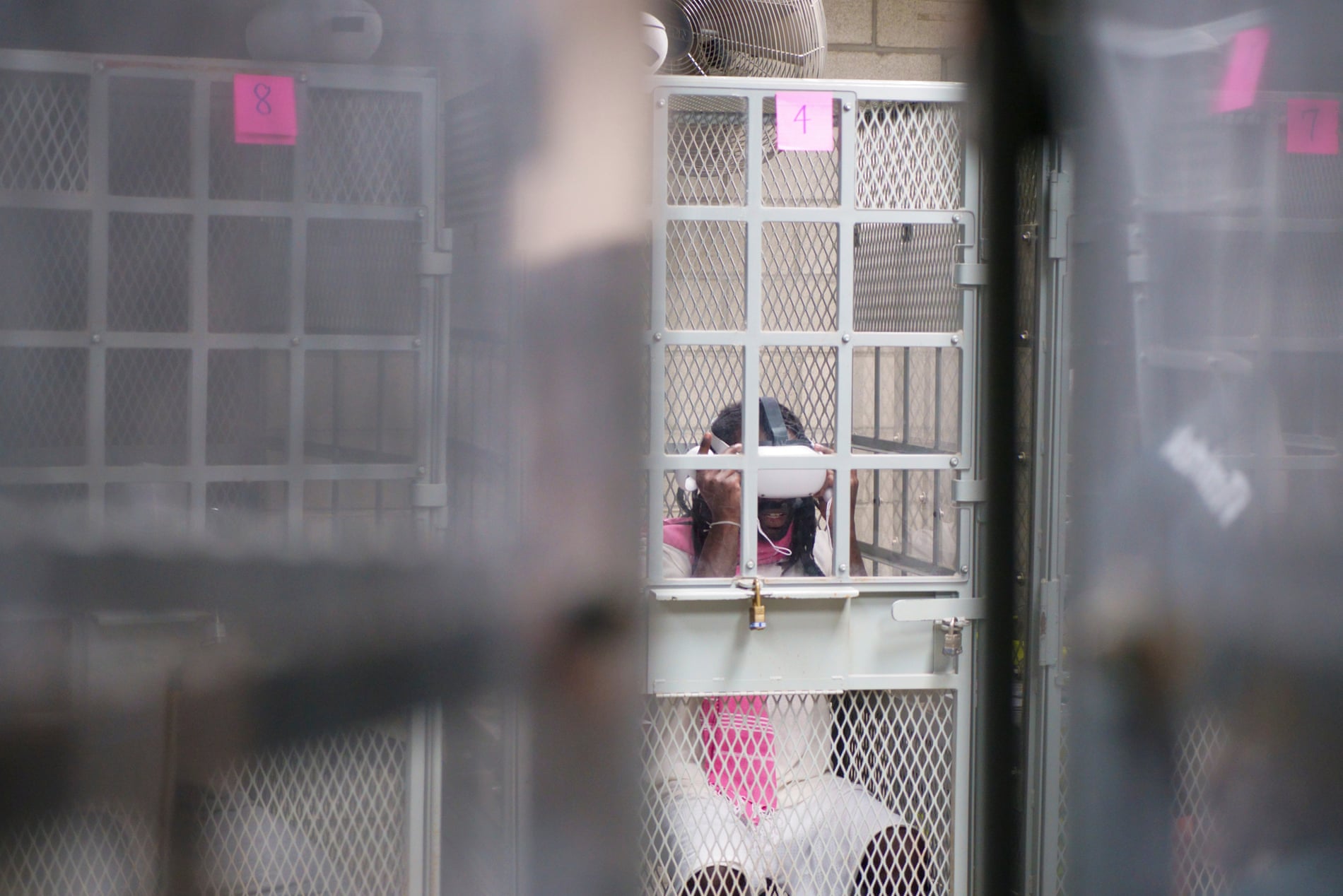 Persona dentro de una celda de malla metálica con una puerta cerrada con candado. Lleva un visor de realidad virtual y sostiene el dispositivo con ambas manos. La celda está etiquetada con el número '4' en un papel rosa. En el fondo, se observan otras celdas similares en un entorno industrial con ventiladores en la parte superior.