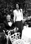 Oates in a white lacy blouse and dark trousers stands beside Smith who is sitting on a chair in black tie in what looks like a garden.