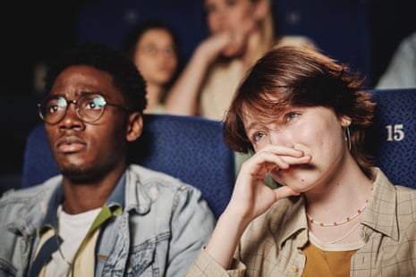 A woman with her hand near her face appears emotional while watching a movie in a cinema, with a man beside her.