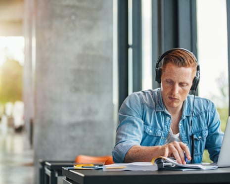 University student working on academic assignment<br>Portrait of young student wearing headphones sitting at the table in library and reading book. University student finding information for his academic assignment.