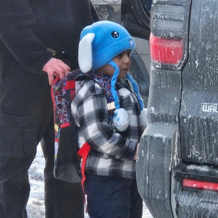 A boy wearing a hat faces a vehicle with a person grasping his backpack.