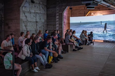 Visitors sit on benches and watch a screen