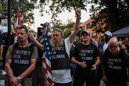 Grim-looking group of white people wearing black T-shirts with white lettering that says Protect Our Children, men looking grizzled with long beards, wearing baseball caps and carrying American flags.