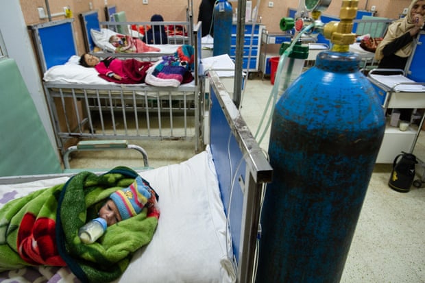 An infant swaddled in a blanket lies on a bed in a hospital ward sucking on a milk bottle with other small children on other beds 