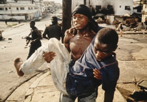 A NPFL Patriotic Front fighter carries a wounded boy comrade as soldiers with guns stand in the background