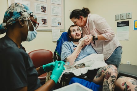 A woman kisses a man's head while he sits at a doctor's appointment, while a nurse refills medicine into a pump